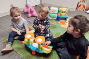 mitchell-infant-toddler-1-600×400 Mitchell group of three toddler girls playing with a colorful music instrument with toys in the background